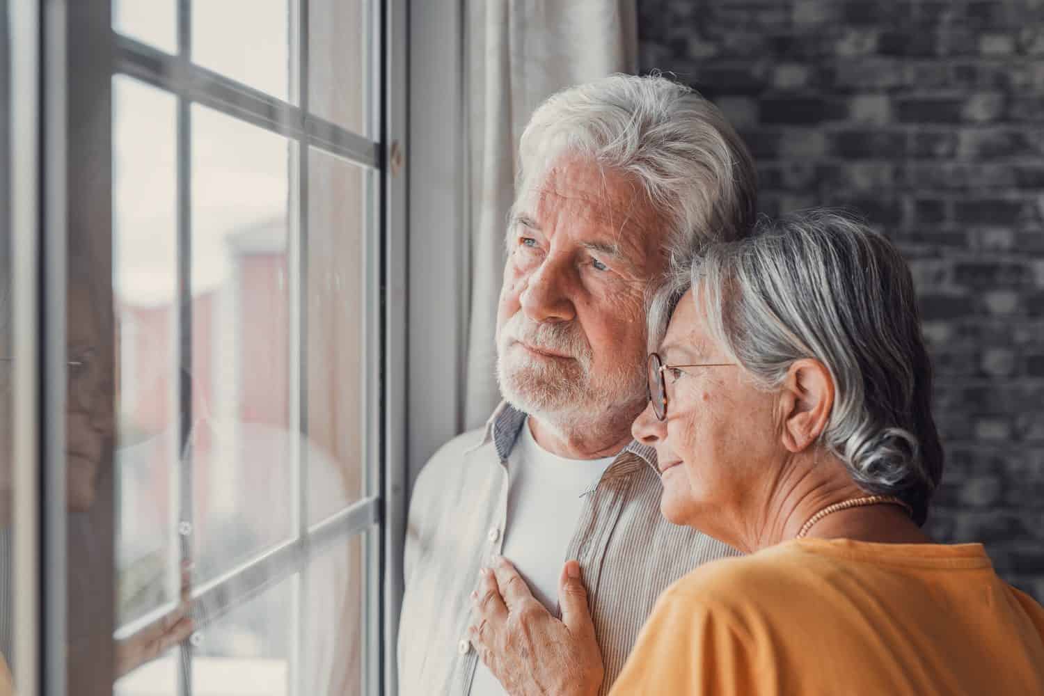 Thoughtful mature aged man with eyeglasses looking into the distance out the window, thinking about personal problems. Old wife comforting and hugging sad husband, copy space