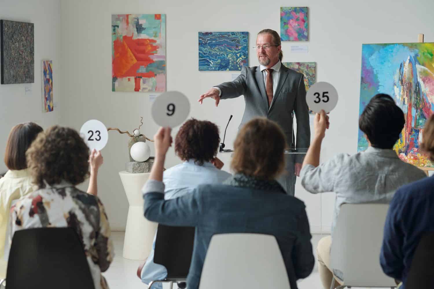 Group of people holding up tablets with numbers up while sitting at auction in art gallery