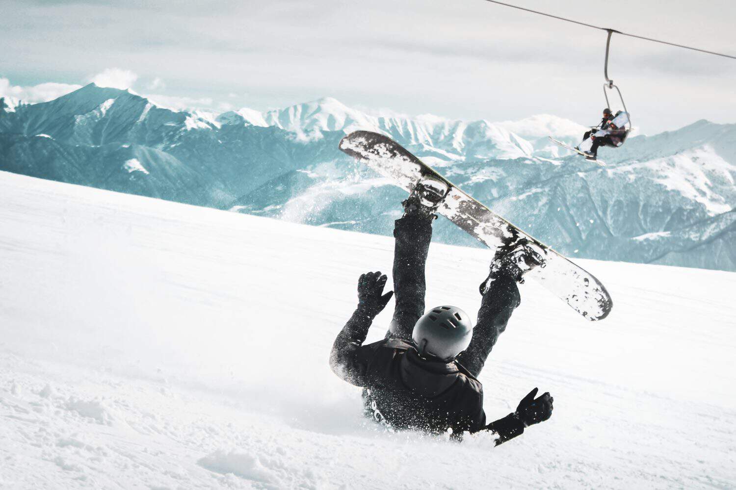 Snowboarder painfully falling backwards with snow splashes on snowy off-piste ski slope and old chairlift in background. Injured skier. The concept of trauma. Impatient bad luck Image in black and white tones.