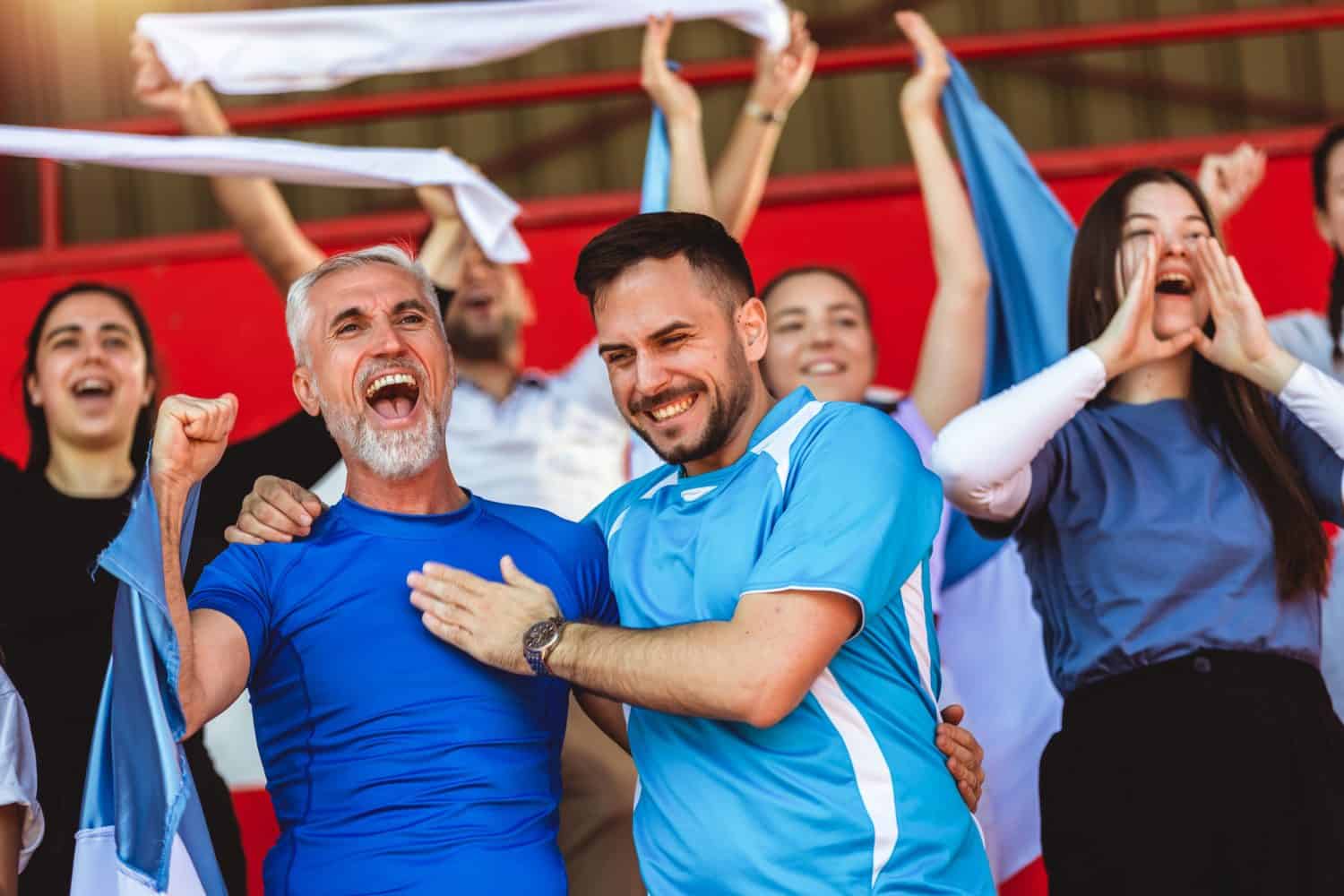 Sport fans cheering at the game on stadium. Wearing blue and white colors to support their team. Celebrating with flags and scarfs.