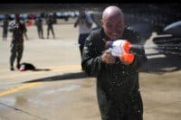 A bald man in a dark green flight suit smiles broadly with his eyes closed, holding a white and orange Super Soaker water gun that sprays water forward. Water droplets splash around him and the ground, indicating a playful water fight. The background is blurred, showing other figures and parts of an aircraft on a tarmac under sunlight.