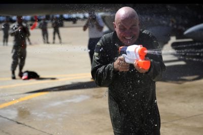 A bald man in a dark green flight suit smiles broadly with his eyes closed, holding a white and orange Super Soaker water gun that sprays water forward. Water droplets splash around him and the ground, indicating a playful water fight. The background is blurred, showing other figures and parts of an aircraft on a tarmac under sunlight.
