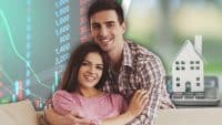 A smiling couple, a man embracing a woman, sits on a couch. Behind them, a translucent overlay displays a financial stock chart with green lines and red and green candlestick patterns, along with numbers. To the right, a white miniature house model sits on a grey calculator.