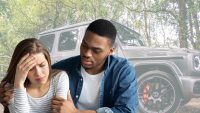 A young woman with brown hair, wearing a striped shirt, holds her hand to her forehead in distress. A young man with dark skin and a denim shirt comforts her with his arm around her shoulder. Behind them, a gray Mercedes G-Wagen SUV is partially visible, blurred amidst green trees.