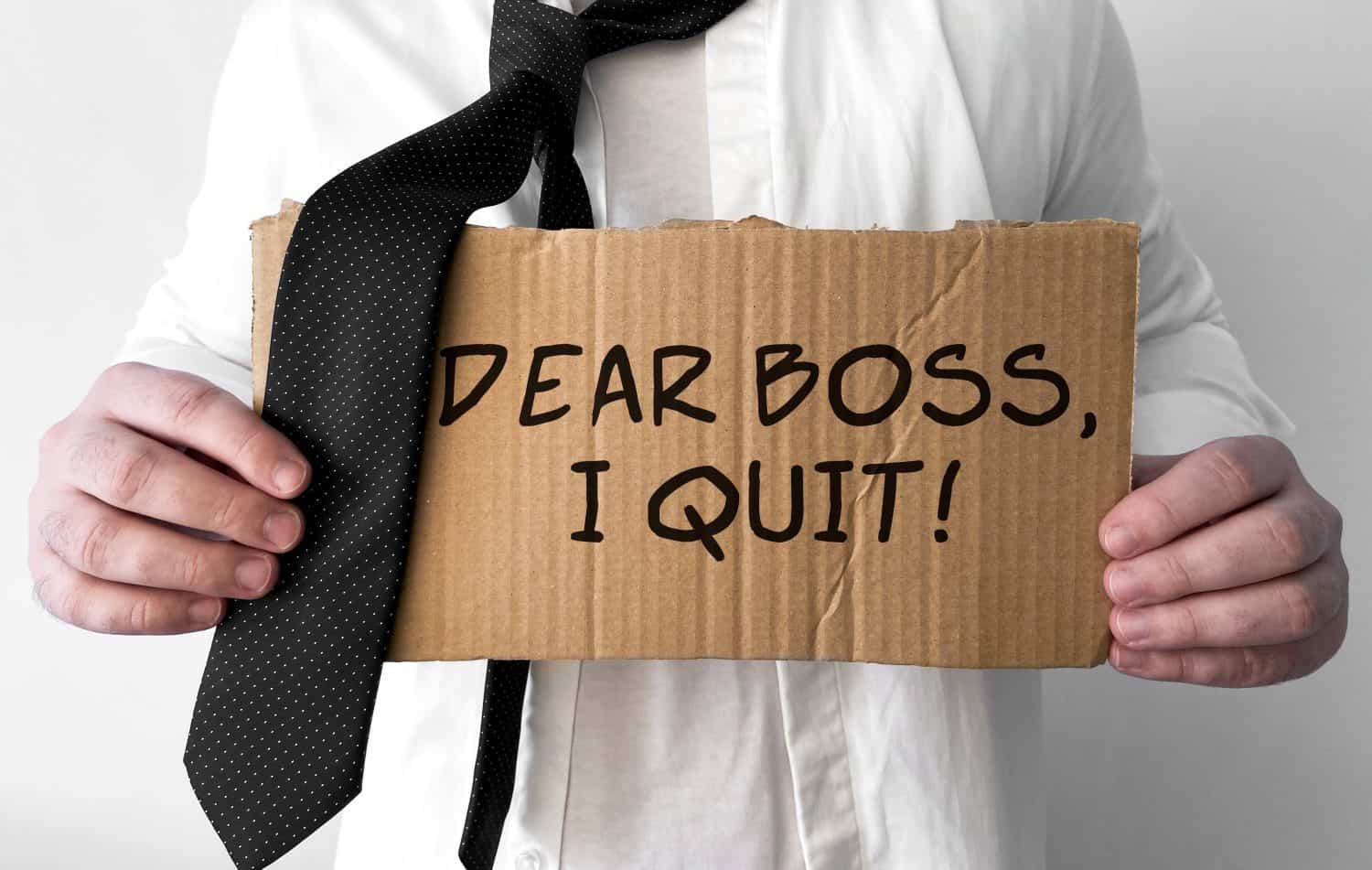 A disheveled business man holds up a cardboard sign with the words Dear Boss I Quit