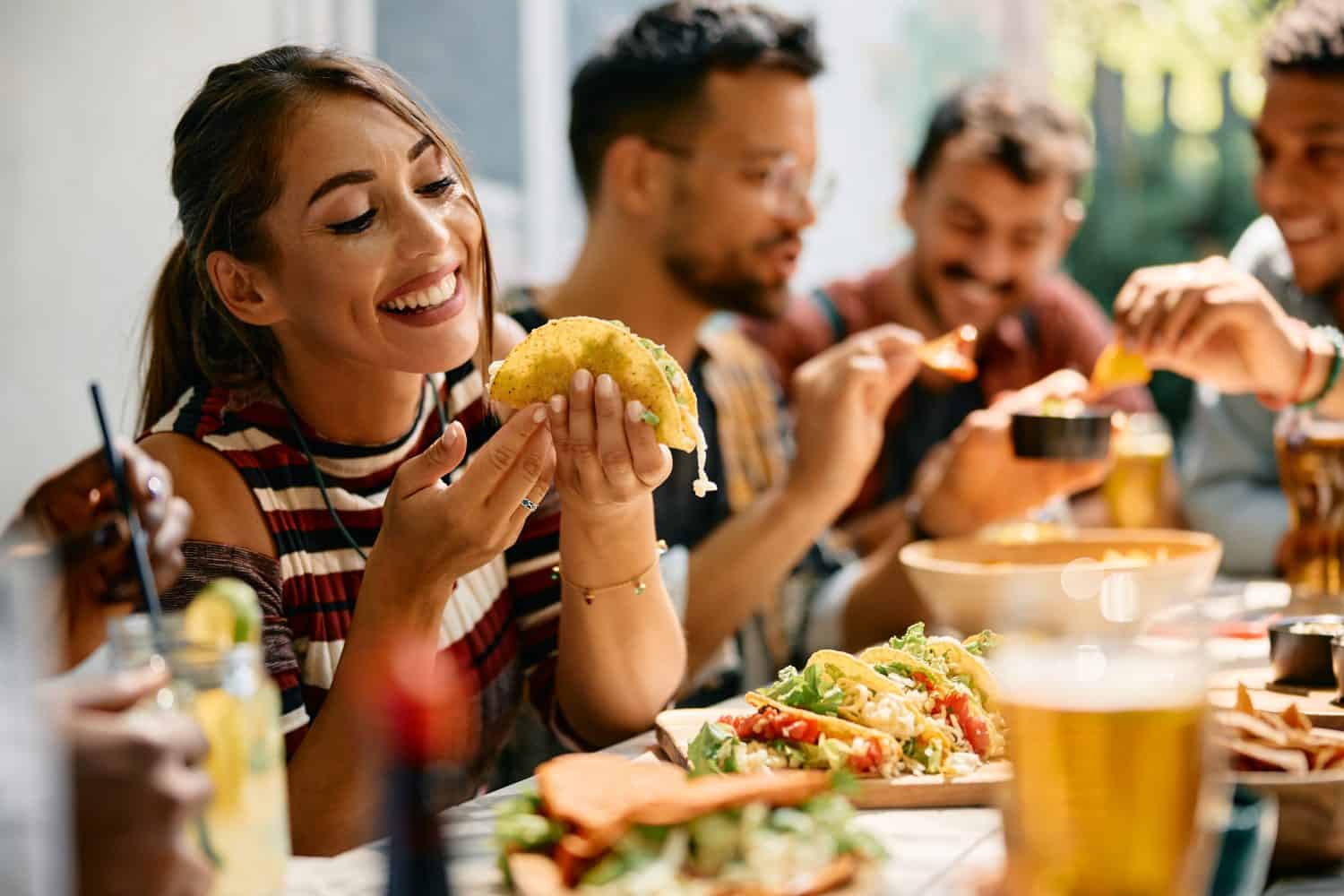 Young woman enjoying a taste of tacos while having lunch with friends in the restaurant.