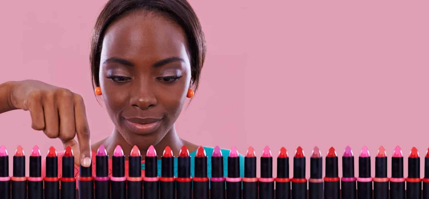 Lipstick, makeup and color choice with black woman in studio isolated on pink cosmetic background. Make, retail and shop for beauty products with a young customer on decision space