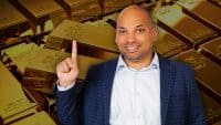 A smiling man with a bald head and light brown skin, wearing a light blue collared shirt and a dark blue checkered blazer, points his right index finger upwards. He is positioned in front of a blurred background filled with numerous shiny gold bars, some showing text like 'FINE GOLD' and 'NET WT 1000g'.
