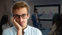 A young man with light brown hair, black-rimmed glasses, and a light blue collared shirt looks at the viewer with a bored expression, resting his chin on his hand. Behind him, a blurred image shows a man in a blue suit presenting a bar chart on a large screen to an audience in a modern conference room.