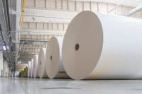 A low-angle view shows a long line of very large white paper rolls, appearing as light-colored cylinders, neatly arranged on a reflective grey concrete floor inside a vast industrial building. The rolls extend into the distance, with the nearest ones prominently displaying their dark circular core. Overhead, the ceiling features visible industrial beams, yellow and grey ductwork, and hanging fluorescent and dome light fixtures, suggesting a large-scale manufacturing or storage facility.