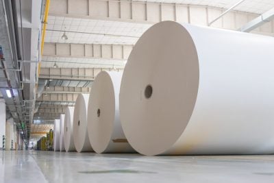 A low-angle view shows a long line of very large white paper rolls, appearing as light-colored cylinders, neatly arranged on a reflective grey concrete floor inside a vast industrial building. The rolls extend into the distance, with the nearest ones prominently displaying their dark circular core. Overhead, the ceiling features visible industrial beams, yellow and grey ductwork, and hanging fluorescent and dome light fixtures, suggesting a large-scale manufacturing or storage facility.