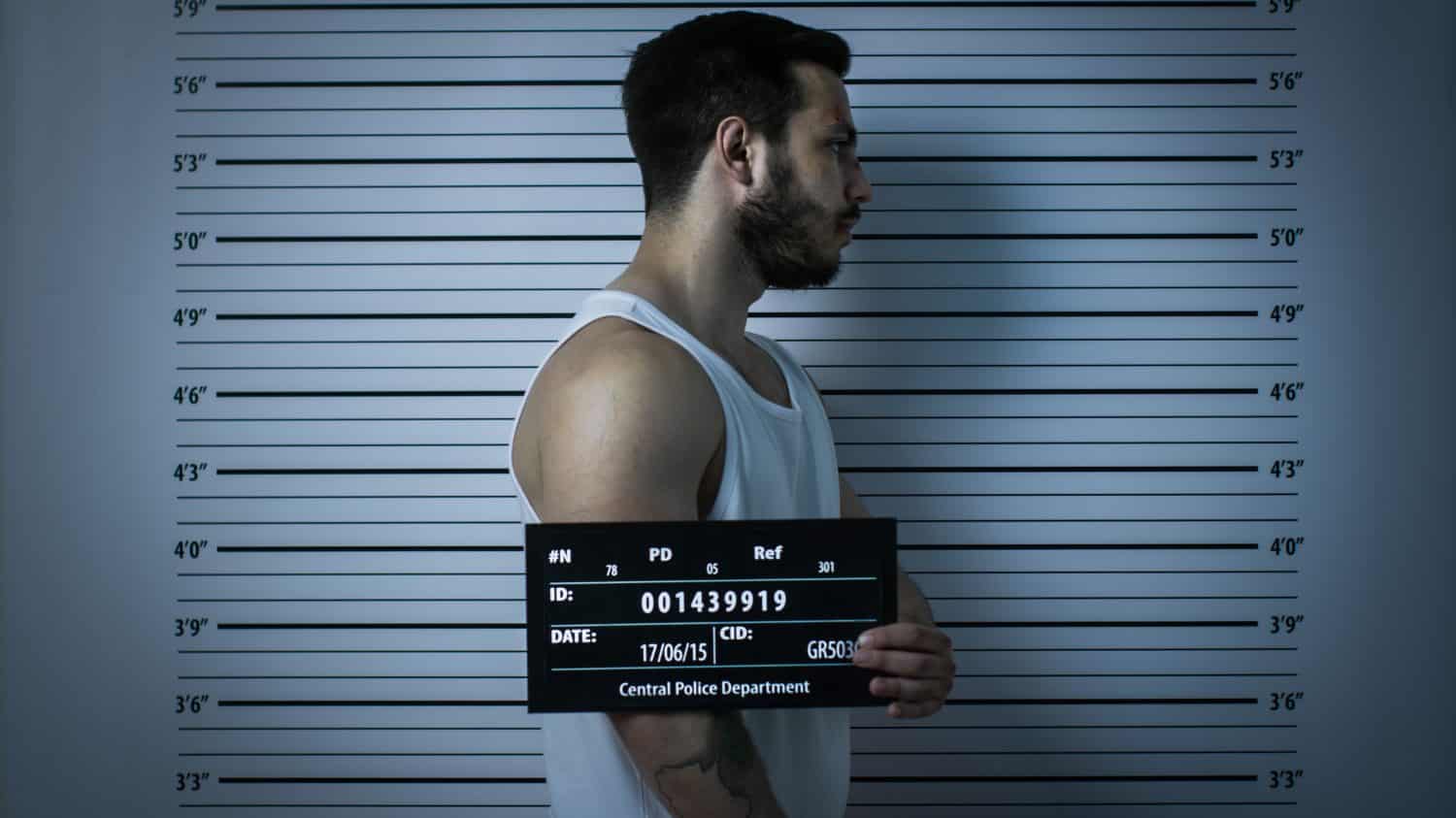 In a police station, a beaten man poses for a side-view mugshot. He is wearing a shirt, badly bruised and holding a placard. Elevation graph in the background. Shot with dark, cold lights, vignette filter