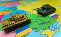 A close-up shot of a world map showing Eastern Europe, with Ukraine prominently visible. Two miniature toy tanks are positioned on the map, facing each other. A gold-colored tank sits on the region of Belarus, adjacent to a small Ukrainian flag. A dark green tank is on Ukraine, near a small Russian flag. The map details cities like Dnipropetrovsk and the Sea of Azov.