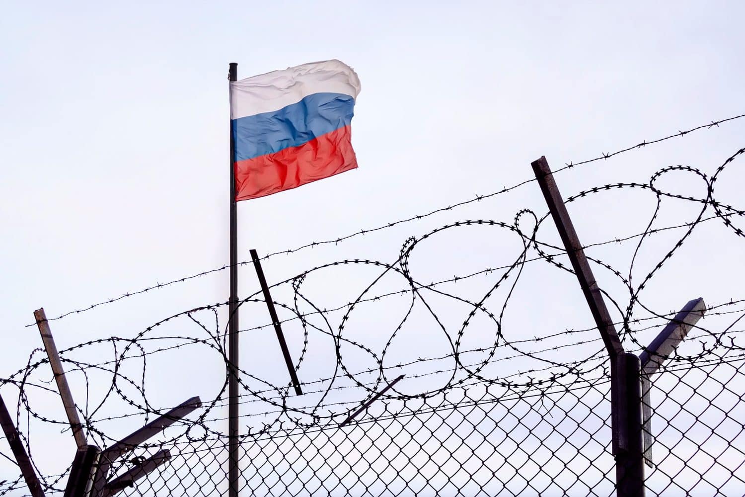 View of the Russian flag behind barbed wire against cloudy sky. Concept of anti-Russian sanctions. A border post on the border of Russia. cancel culture Russia in the world