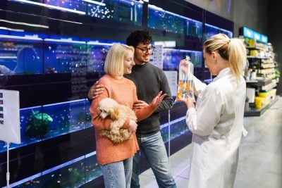 A couple and a blonde female store employee stand in a pet store aisle lined with glowing blue-lit aquariums. The blonde woman, wearing a white lab coat, holds up a clear plastic bag containing two orange goldfish for the couple. The woman in the couple, with short blonde hair and an orange sweater, holds a small light brown fluffy dog in her arms and smiles at the goldfish. The man in the couple, with dark curly hair and glasses, has his arm around her and also smiles. The background shows shelves of pet supplies.
