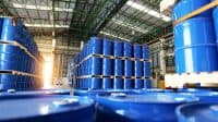Large blue industrial barrels are stacked high on wooden pallets inside a sprawling warehouse. Rows of barrels extend into the background, where a bright light source creates a lens flare effect. The warehouse features an industrial ceiling with metal beams and overhead lighting fixtures.