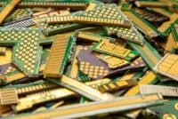 A close-up, top-down view of a large, messy pile of electronic waste. The pile consists of numerous small, irregular fragments of green circuit boards with visible gold-plated contacts and traces. Some pieces are rectangular, others triangular or square, showing arrays of gold circles and long gold strips. The dominant colors are vibrant green, shiny gold, and dark grey/black on some component bases, indicating a mix of materials.