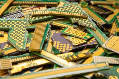 A close-up, top-down view of a large, messy pile of electronic waste. The pile consists of numerous small, irregular fragments of green circuit boards with visible gold-plated contacts and traces. Some pieces are rectangular, others triangular or square, showing arrays of gold circles and long gold strips. The dominant colors are vibrant green, shiny gold, and dark grey/black on some component bases, indicating a mix of materials.