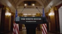 A front-on view of a black podium with the words 'THE WHITE HOUSE' in white text above a thin red line, and 'WASHINGTON' in white text below. Two microphones extend from the top of the podium. American flags are partially visible on both sides of the podium. The background is an ornate, light-colored room with columns and sconce lighting, suggesting a formal government setting.