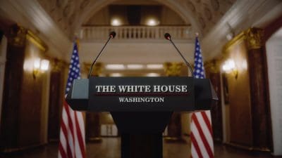 A front-on view of a black podium with the words 'THE WHITE HOUSE' in white text above a thin red line, and 'WASHINGTON' in white text below. Two microphones extend from the top of the podium. American flags are partially visible on both sides of the podium. The background is an ornate, light-colored room with columns and sconce lighting, suggesting a formal government setting.