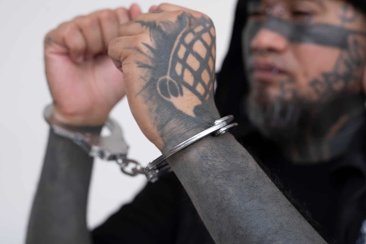 An Asian man with densely inked facial tattoos, looking sideways in handcuffs, against a plain white background. A convicted felon who has been arrested. Judged and prejudiced by his appearance.