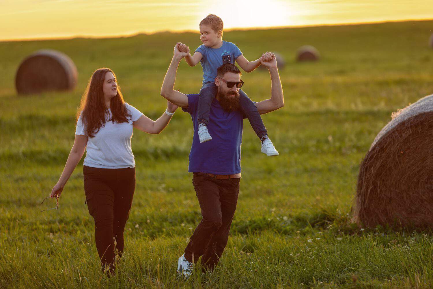 A young family with their little boy on their father's shoulders, running on a hilly meadow in the background of the sunset.