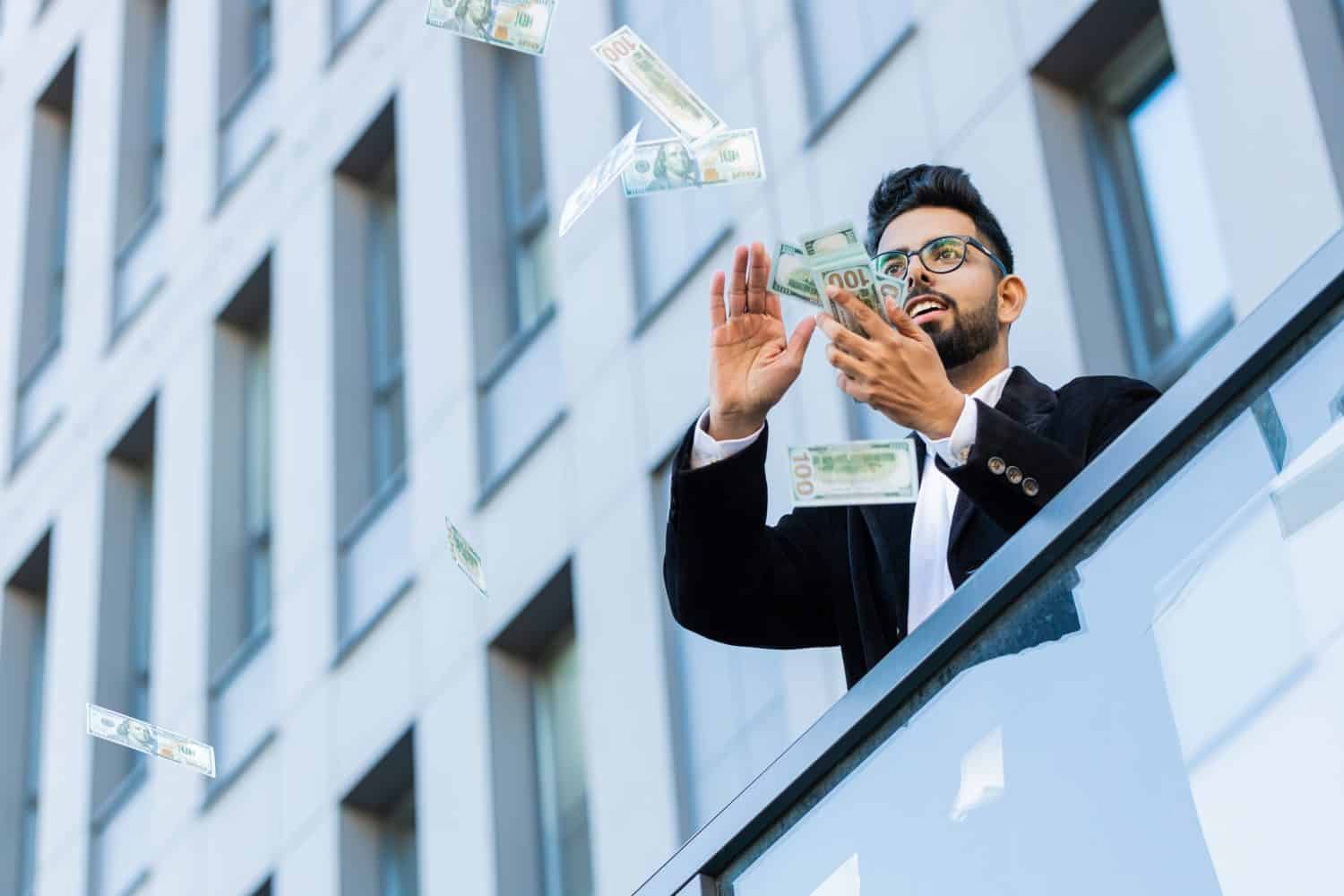 Happy rich Indian businessman wasting money, dollars in cash, around earnings, big profit, win lottery, share, celebrate, charity donation outdoors. An Arab Hindu freelancer in the city center