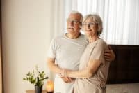 Smiling senior couple in love hugging at nursing home and looking through window.