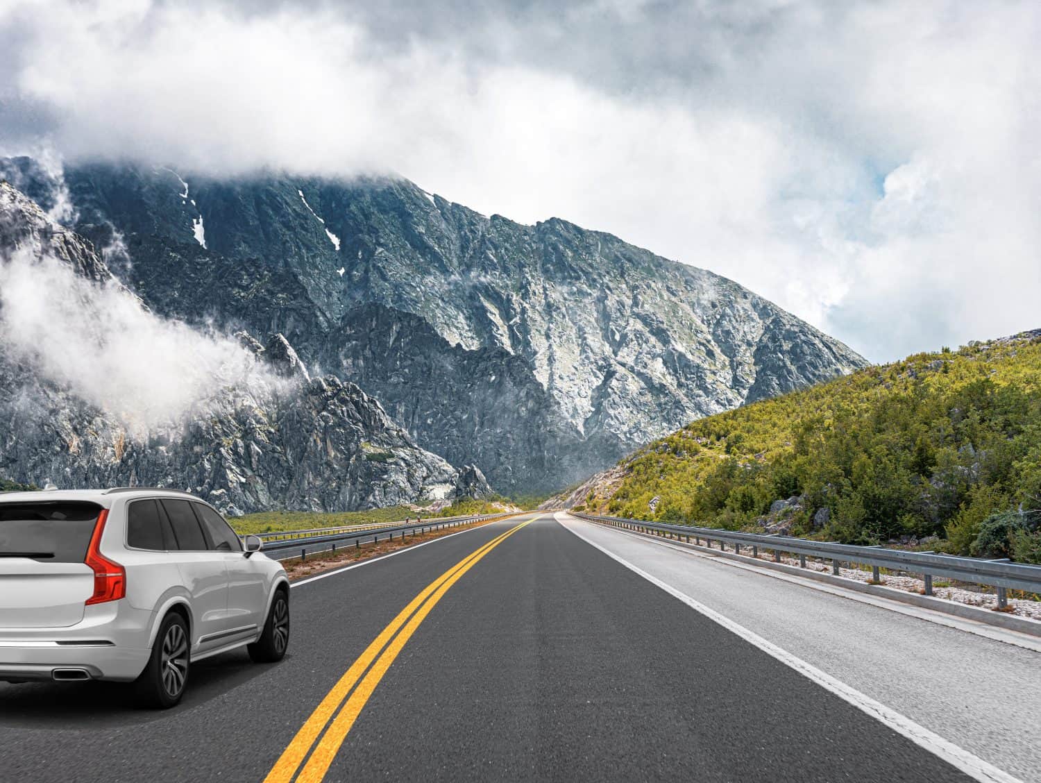 A white car drives along the highway against the backdrop of rocky mountains on a sunny day.