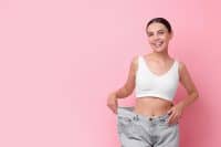 Diet and weight loss concept. Happy young woman in big jeans showing her slim body on pink background. Space for text