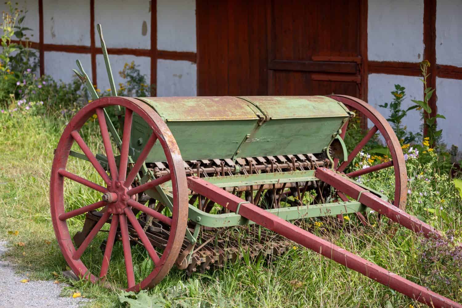 An old fashioned green and red seeder resting on the grass with a rustic wooden building in the background. The seeder has large wooden wheels and a metal mechanism for planting seeds.