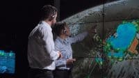 Two individuals, a man and a woman, both wearing headsets, are standing in a dimly lit control room. The woman on the right points at a large multi-panel screen displaying an Earth map with vivid blue, green, and orange weather or environmental data overlays. The man on the left looks towards the screen, next to her.