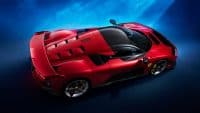 An aerial view of a glossy red Ferrari sports car from the rear three-quarter angle. The car has a prominent integrated rear spoiler, a black roof and engine cover with vents, and black multi-spoke wheels with yellow brake calipers. The background is a gradient from bright blue sky at the top to a dark, speckled blue surface below, creating a dramatic contrast with the car's vibrant red.