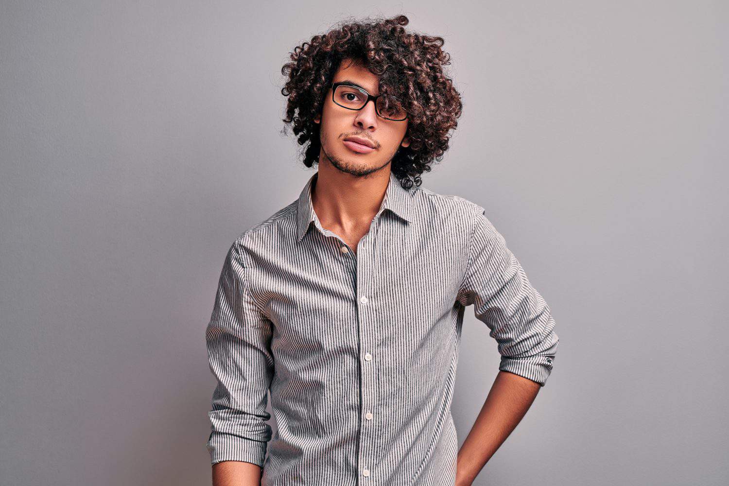 Young man in stylish eyeglasses seriously looking at camera standing on isolated gray background. Confident arabian handsome boy. Studio shot
