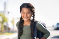 Portrait of happy indian girl with school bag standing outdoor. Little happy schoolgirl going to school, back to school, education concept. Cute mixed race female student smiling and looking at camera