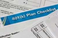 A blue and white banner with '401(k) Plan Checklist' in bold white text is diagonally placed over several white financial documents. Portions of the underlying forms are visible, showing printed text, dotted lines, dollar signs, and sections labeled 'SIGN HERE' and 'Signature of employee'. The papers are white with black text and lines.