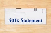 A white standard-sized envelope with the words '401k Statement' printed in large, bold dark blue font on its front. The envelope features a transparent rectangular window in the upper left section for an address and various black lines and a square in the top corners, indicating postal markings. It rests on a light brown wooden surface, showing natural wood grain patterns.