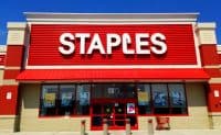 The vibrant red and beige exterior of a Staples store on a sunny day. The large white 'STAPLES' logo is prominently displayed on the red corrugated facade, which is topped with a white trim. A red awning extends over the entrance and large windows, which display promotional signs for 'Clearance' and 'Ship it here. UPS'. Concrete bollards line the front sidewalk, and a red shopping cart is visible to the right. A 'NO PARKING FIRE LANE' sign is posted on a beige pillar to the left.