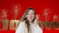 A smiling woman with long wavy blonde hair, wearing a cream cable-knit sweater, looks directly at the viewer. Golden confetti showers around large, metallic gold numbers that spell '100,000', set against a solid bright red background. The woman's left hand is raised to her ear, and her expression is one of pure joy and excitement.