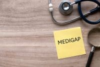 A yellow sticky note with 'MEDIGAP' written in black capital letters lies on a light brown wooden surface. A silver and blue stethoscope is coiled at the top right, and a silver magnifying glass with a black handle is positioned to the right of the note.