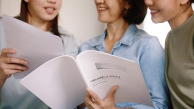 A close-up view of three Asian women looking at an open white folder titled 'INSURANCE POLICY'. The woman in the center, wearing a light blue denim shirt, holds the document. A gold ring is visible on her right ring finger. The women appear engaged and thoughtful, focusing on the contents of the policy.