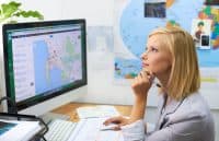 A blonde woman in a grey blazer is seated at a desk, looking up and to the right thoughtfully while holding a pen to her chin. In front of her is a computer monitor displaying a map application with many location pins. Her right hand is on a white computer mouse. On the wall behind her is a large world map with smaller images pinned to it. A green plant is visible on the left side of the desk.