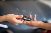 Two fair-skinned hands are shown in close-up, exchanging a sleek, black car key fob with silver buttons. The left hand extends the key towards the right hand, which is open and ready to receive it. The background is a blurred, dark gray with soft circular light reflections.