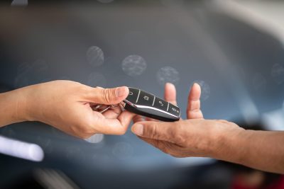Two fair-skinned hands are shown in close-up, exchanging a sleek, black car key fob with silver buttons. The left hand extends the key towards the right hand, which is open and ready to receive it. The background is a blurred, dark gray with soft circular light reflections.