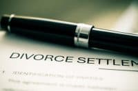 A black pen with a shiny silver band rests diagonally on a white document. The document's visible text includes the bold, capitalized words 'DIVORCE SETTLEMENT' and '1. IDENTIFICATION OF PARTIES' below it. The background is softly blurred.