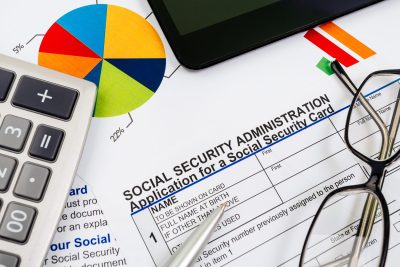 An overhead shot of a Social Security Administration application form, partially obscured by a silver calculator on the left, a silver pen, and black-rimmed eyeglasses on the right. In the background, a colorful pie chart with visible percentages of 5% and 22% indicates financial data.