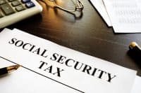 A close-up shot of a white document with 'SOCIAL SECURITY TAX' printed in large, bold, black capital letters. The document rests on a dark wooden desk. To the upper left, a white calculator with grey and blue keys is visible. A pair of gold-rimmed reading glasses lies above the document, and a black and gold fountain pen is positioned at the bottom left, pointing towards the text. Other financial papers with columns of numbers are subtly visible in the background.