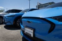 A close-up view of two bright blue Ford Mustang Mach-E electric vehicles parked outdoors at a dealership. The front vehicle displays a "Mustang" pony logo and a "Fox Motors" license plate frame. In the background, a white Lincoln Aviator and a modern Ford dealership building with the Ford logo are visible under a clear blue sky, suggesting a sunny day.