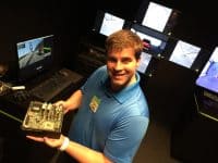 A smiling man in a blue polo shirt holds up an NVIDIA Drive CX circuit board, revealing its green motherboard and various electronic components. Behind him, multiple monitors are arranged, displaying screens of a driving simulation, including a view of a bridge, a white van on a road, and different virtual camera angles of a red car, all glowing in a dimly lit room.