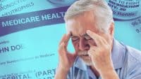 An older man with gray hair and a beard, wearing a light blue shirt, holding his temples as if he has a headache. His eyes are closed, indicating discomfort or stress. The background is a composite of blurred Medicare documents and prescription labels, all bathed in a strong blue and teal filter, emphasizing the theme of healthcare and financial worries.